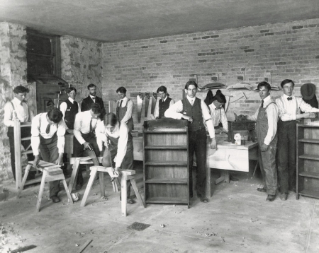 Posed image of students in a cabinetmaking class. At the center of the image a student leans against a finished cabinet. Other students are engaged in various activities like sawing and planing wood. A teacher observes in the background.