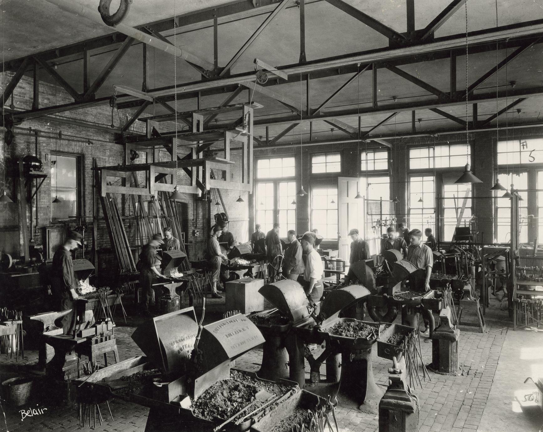 A interior scene of a classroom foundry. Dozens of small forges are arranged in rows. Students are working on various wrought iron projects.