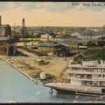 A colorized postcard of the ship yards at Manitowoc. At the far left there is a grain elevator and large cargo ship. Also visible are many warehouses and large shop buildings, and a ship in scaffolding wither being built or repaired.