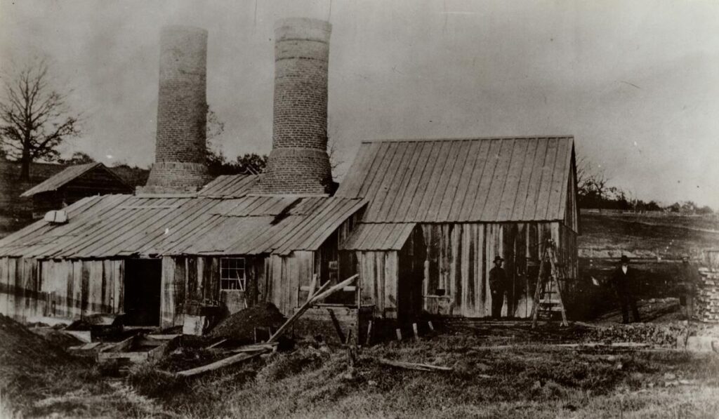 A period photograph of a open-hearth furnace, wood sheathed building with a pair of large round chimneys.