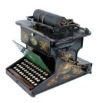 A metal typewriter with keyboard lid. The metal is ornamented with floral designs.