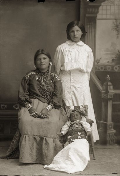 Full-length studio portrait in front of a painted backdrop of two young Ho-Chunk women and a Ho-Chunk infant. The woman sitting on the left is wearing regalia, and the woman standing on the right is wearing contemporary clothing. The infant is in a cradleboard on the floor between the two women.