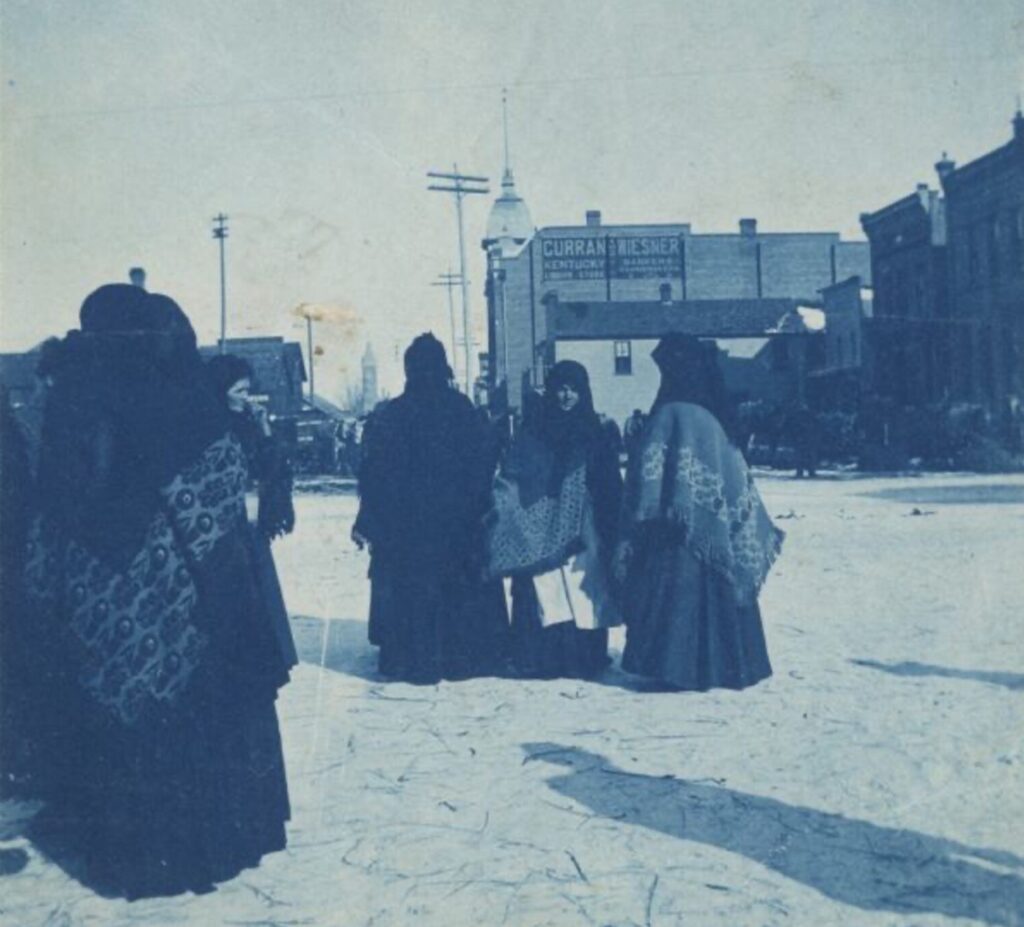 Polish immigrant women at the Market Square wearing traditional, national clothing. In the background are brick buildings. A sign on one building reads: "Curran and Wiesner. Kentucky Liquor Store. Bankers, Pawnbrokers."