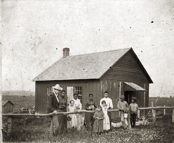 A group of people, posed against a fence in front of a one-room schoolhouse.