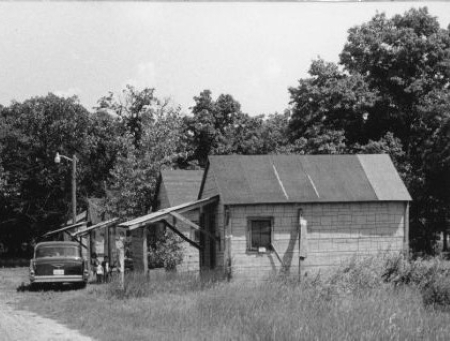 A row of small wooden framed buildings with front porches on posts. The buildings are faced with asphalt siding. At the center, three children are standing in front of a building beside a black car looking into the camera. Farther down the road on the left is a shack and a worker standing by a car across from it.