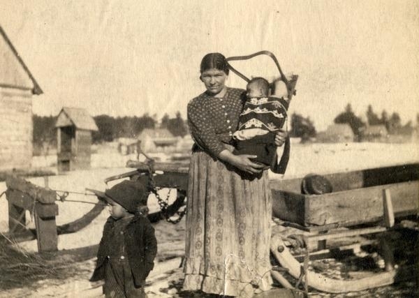 An Lac du Flambeau Ojibwe woman stands outdoors in front of a sleigh in winter. She holds an infant in a cradleboard, another child stands nearby.