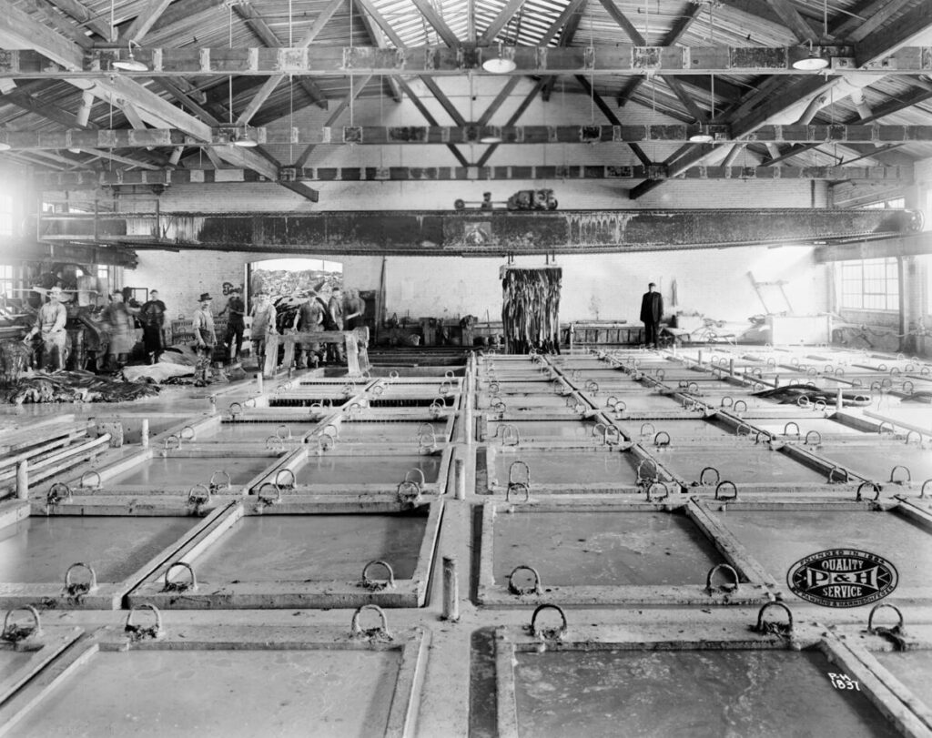View across room of tanks and a Pawling & Harnischfeger type "GN" low overhead crane in a tanning operation. Men are standing on the left towards the back of the building near an open doorway. Hides can be seen piled behind them. Hides are hanging on a rack from the crane near a man on the right. The P&H logo is on the lower right corner and reads "Founded in 1884 By Pawling & Harnischfeger, Quality Service."
