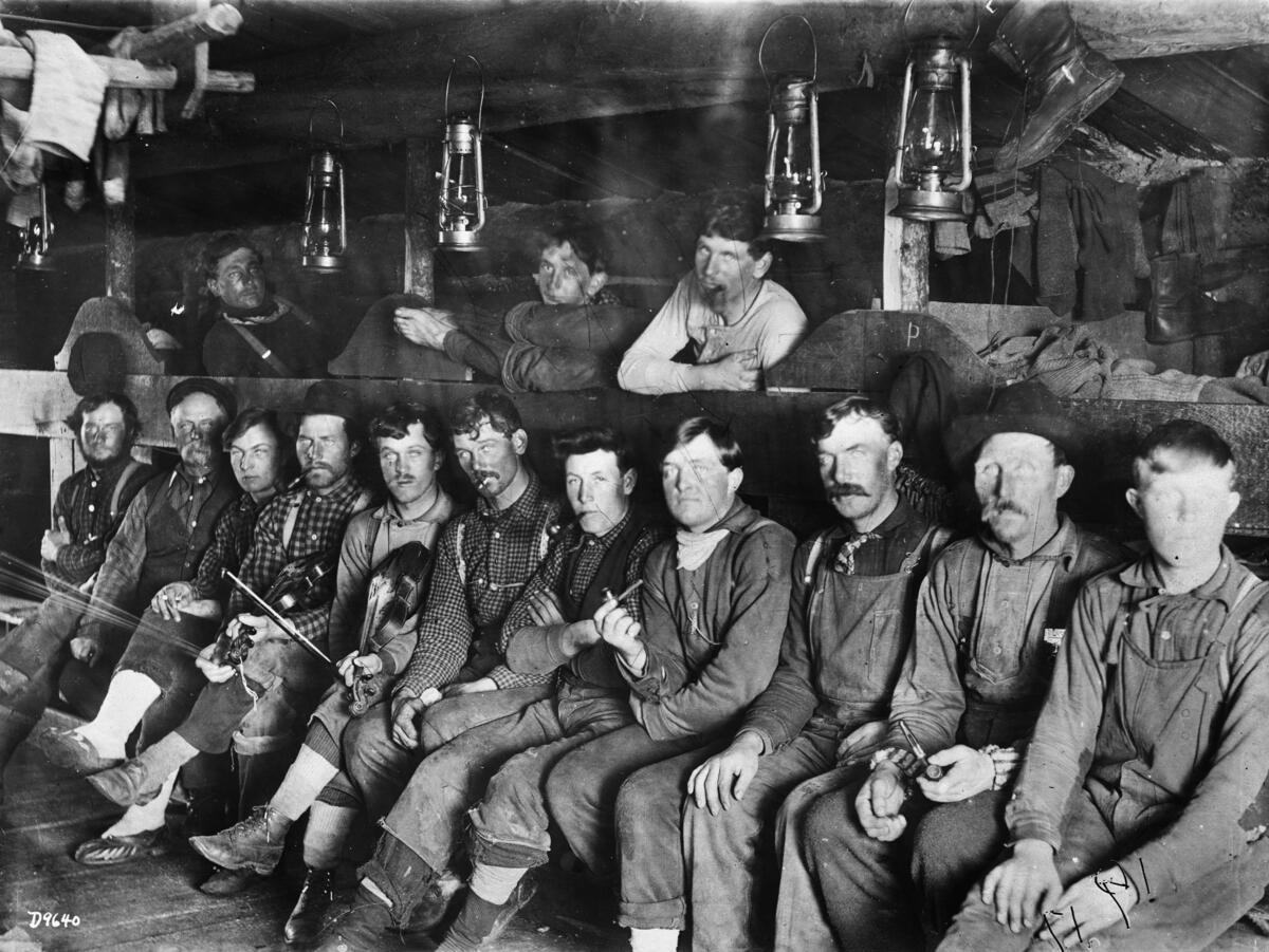 A group of men pose inside a bunk house at a lumber camp building. Some recline on the top bunk, while others sit, many posed with pipes. Two men hold fiddles.