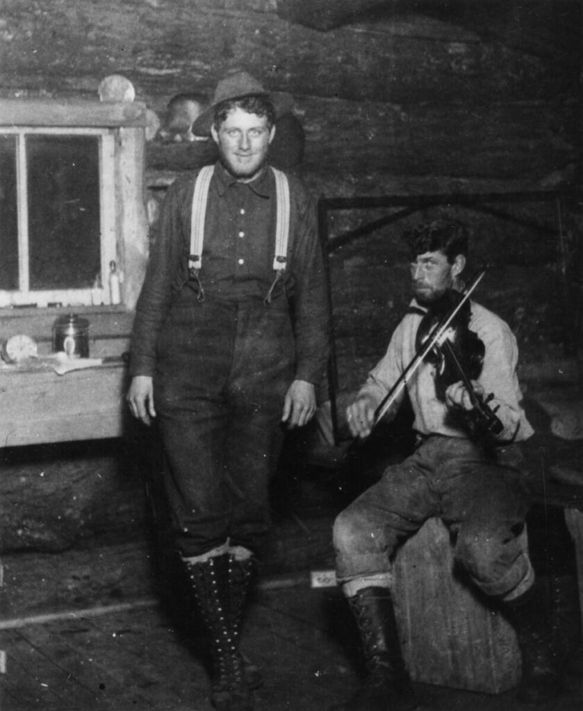 Photo from the interior of a lumber camp building. Two men pose for the camera. One is seated and holding a fiddle to his shoulder, bow across the strings. The other man stands next to him, arms to his side, on the balls of his feet as if he's doing a jig.