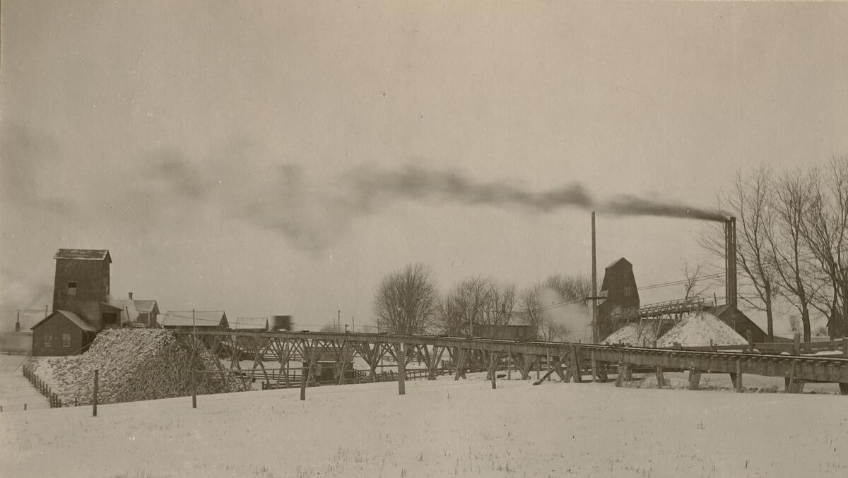 Image of a mine complex, above ground. Image shows hoist houses, long rail ramps for ore carts, and piles of crushed stone.