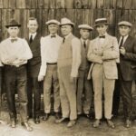An image of seven men in suits posing in front of a barn door.