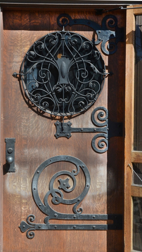 The Wilson Place Mansion door, featuring a circular window with an intricate wrought iron screen of intertwining vines and acanthus leaves around a central cartouche; and very large, strap hinges, also in a vine motif.