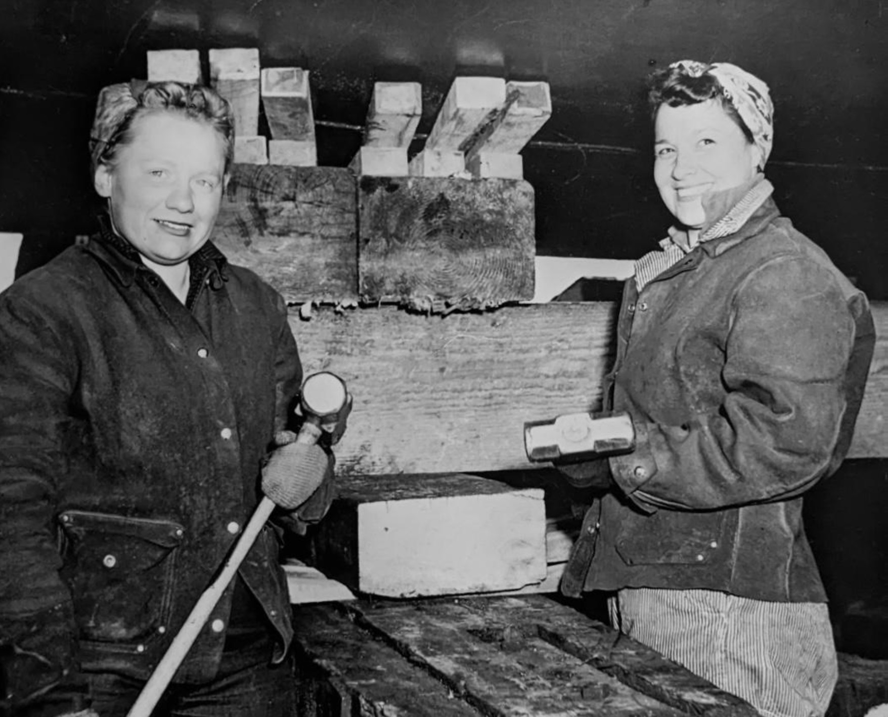Two women pose for the camera. They wear worker’s outfits and hold sledge hammers.