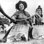 Two women in Swiss costume kneeling in the grass during the Swiss Cheese celebration. One is blowing into an alpine horn while the other holds the instrument.