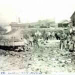 A large group of workers pose for the camera in a port industrial landscape. A large cargo ship in the background. Handwritten caption reads, "1892 Building First Dry Dock."