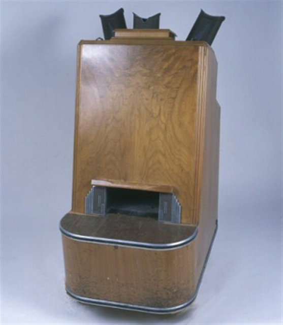 A Simplex fluoroscope machine. A wooden case with a chamber near the bottom for a customer to place their foot. At the top are metal scopes for a clerk to look into the machine.