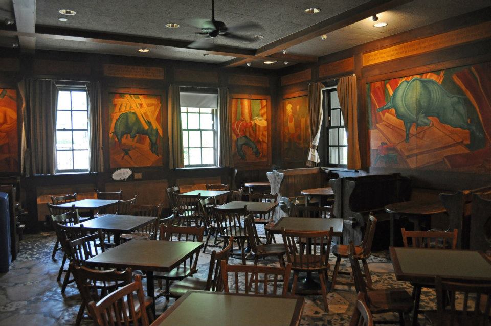 Photograph of a dimly lite room filled with wooden tables and chairs, the walls covered with large colorful murals.