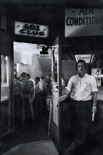 A man holds open a door revealing a bar interior with male patrons standing at the bar.