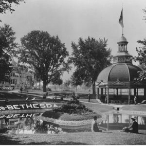 Bethesda Spring Park, ca. 1900. The springhouse stands on the right. The spring runs into the pond, which reflects the words “1868-Bethesda.” The Terrace Hotel can be seen in the left background. People sit on the lawn and stand around the springhouse.
Courtesy of UW Libraries Digital Collections ID: 2004.214.1; 7370