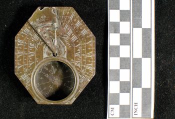 An octagonal, engraved brass sundial and compass. A ruler indicates that it is approximately 8 centimeters (3 inches) long.