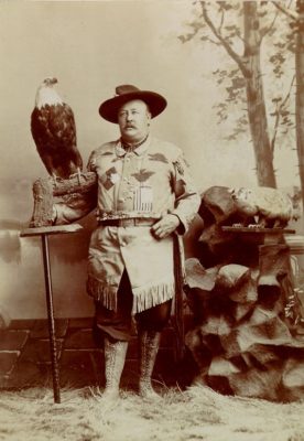 Studio portrait of a man in western costume posed with a live eagle and a mounted badger.