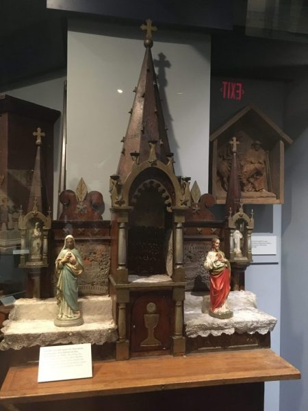 A wooden altar piece, with a tabernacle at the base, and Gothic Style ornamentation including three spires topped with Christian crosses.