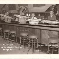 An old postcard showing four men behind a bar holding very large muskies for display