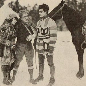 a film still from the 1919 movie, A wilderness trail, showing three people standing before a horse in the woods. One man is wearing a short point blanket coat.