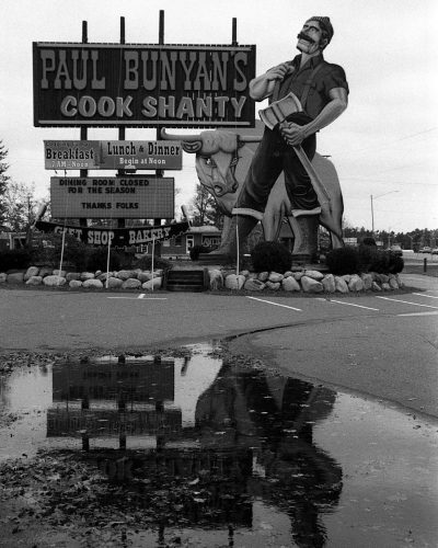 A black-and-white photograph of the sign for Paul Bunyan's Cook Shanty in the Wisconsin Dells, featuring a large scale image of lumberjack Paul Bunyan and his giant blue ox, Babe.