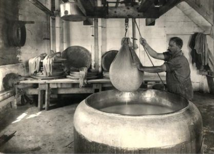 The interior of a cheese factory from 1914 showing a man lifting a sack of curds out of a vat