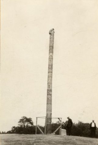 Black-and-white photograph of two men visiting a totem pole. One reads a plaque on a monument at the base of the pole.