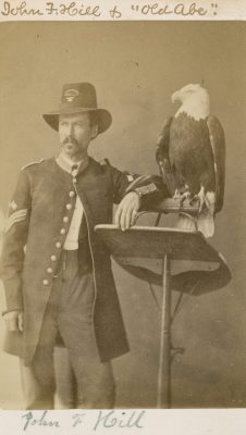 Portrait of a man, in Union Army uniform, next to a bald eagle on a perch. The photo is hand-labeled John F. Hill & Old Abe.