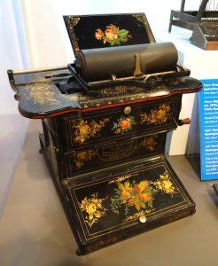 A metal typewriter with closed keyboard lid. The metal is ornamented with floral designs.
