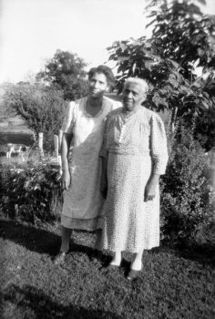 A posed photograph of two women in simple dresses standing in a yard or garden.